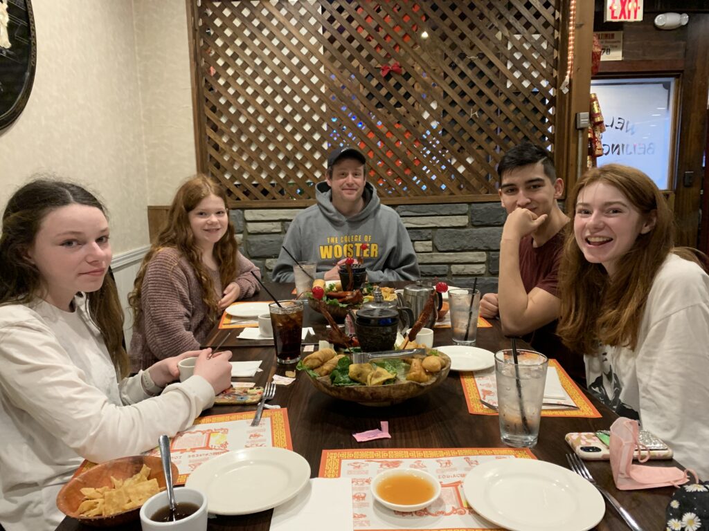 Bri's dad is at the head of the table then Alix and Bri and Bri's two sisters sit on opposite sides. On the table is a platter of traditional Chinese appetizers.