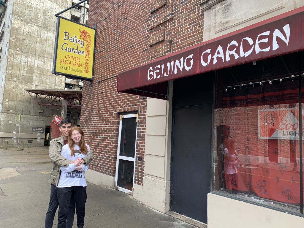 Alix has his arms around Bri and they smile in front of the signage for the Beijing Garden restaurant.