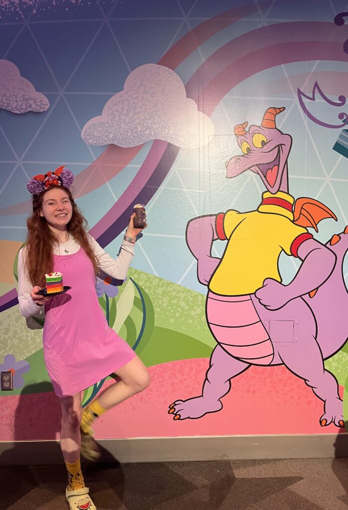Bri holds up a purple drink in a clear cup with Figment's (A purple dragon) face on it in one hand and a rainbow layered cake in the other hand. There is a mural with Figment and a rainbow behind her.