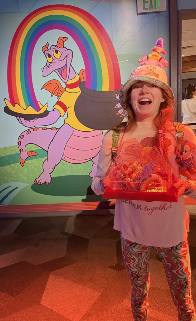 Bri has an open-mouth excited face as she holds up a clear popcorn bucket with figment (a purple dragon) riding a rainbow in it.