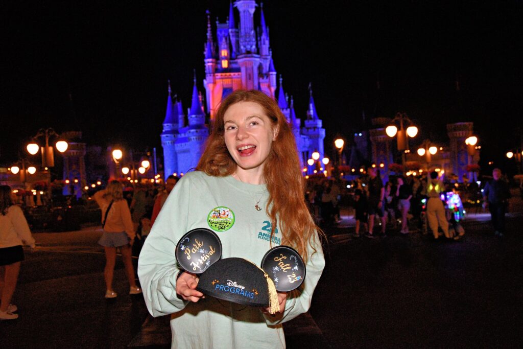 Bri holds her Disney Programs graduation Mickey Ears as she smiles in front of the lit-up Cinderella's castle at night.