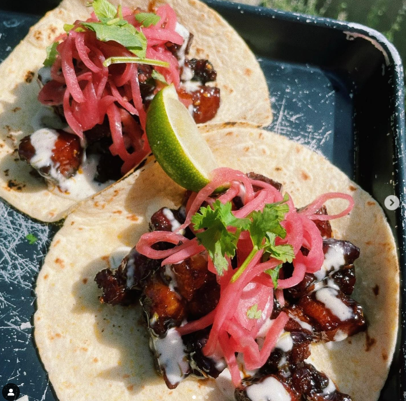 Two pork belly tacos with lime cilantro and pickled red onion layered on a flour tortilla.