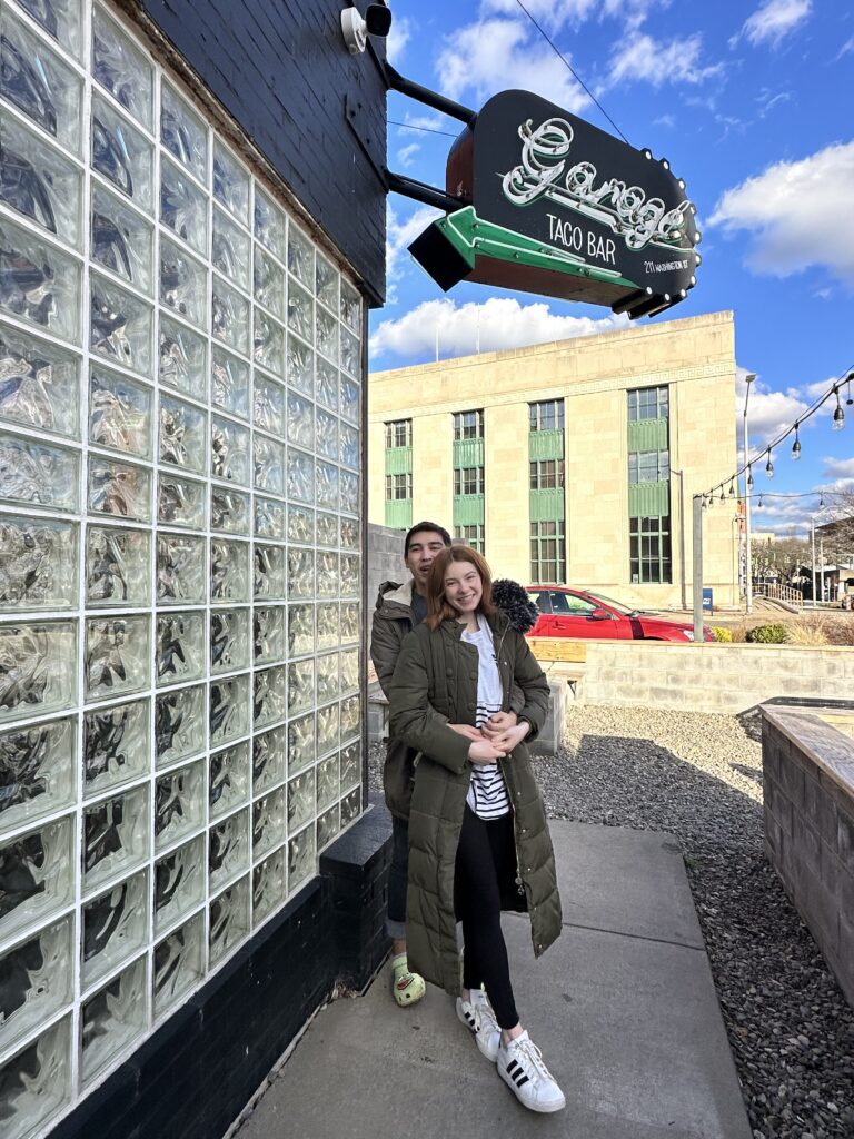 Bri smiles standing in front of Alix who has his arms wrapped around her, The sign above them says "Garage Taco bar" with an arrow pointing to the restauraunt.