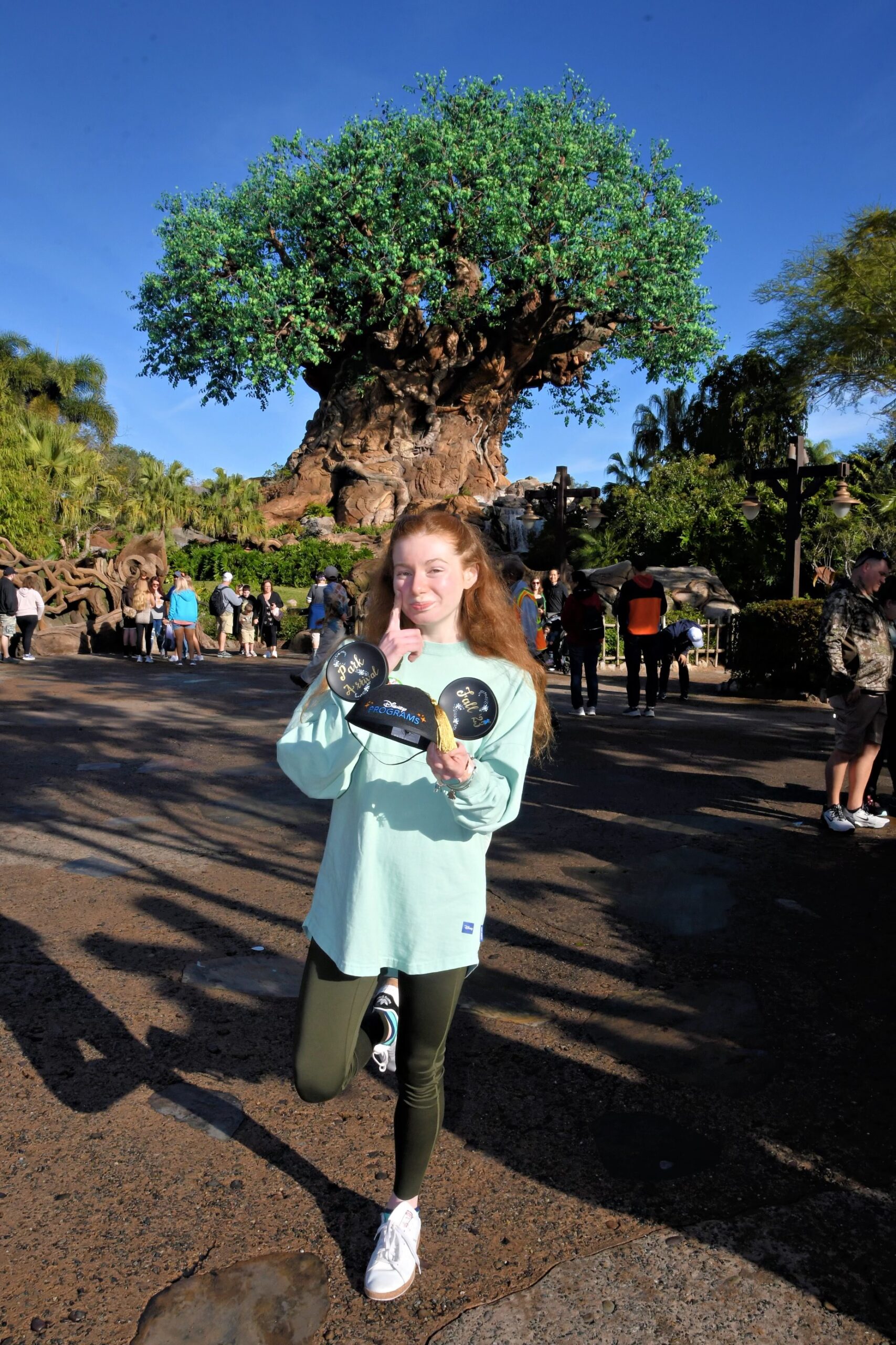 Bri stands in front of Animal Kingdom's tree of life with a finger by her eye imitating a 'sad' face. her other hand holds out a Disney programs mickey ears graduation cap.
