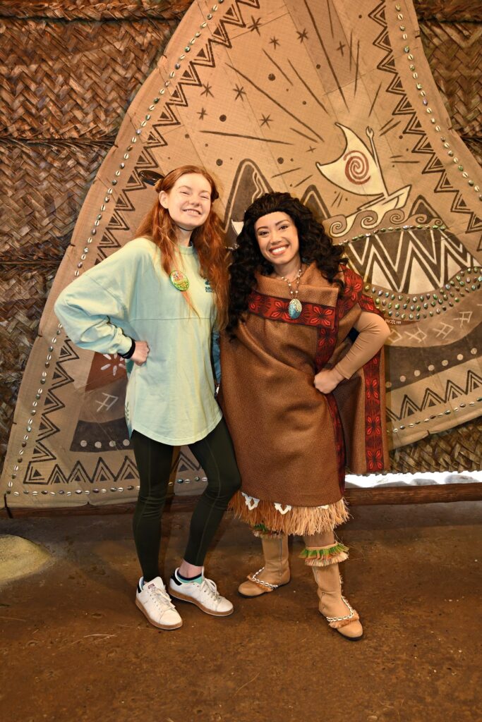 Moana and Bri stand in front of a brown canvas with a wayfinding ship smiling proudly at camera with one hand on their hips and the other arm around each other.