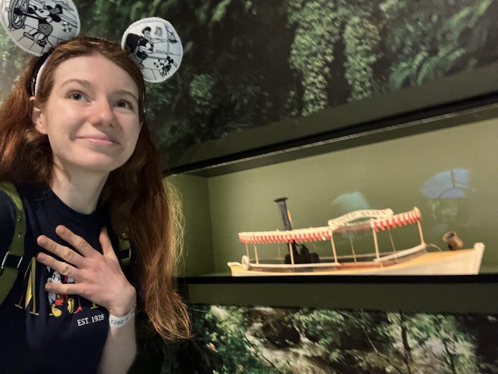 Bri wears steamboat mickey ears and gazes up dreamily with hand over heart as she poses next to a mini jungle cruise boat model.