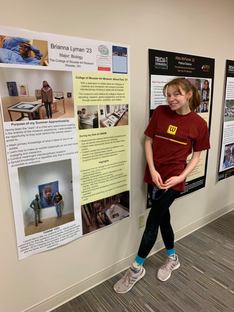 Bri stands smiling next to her museum research apprenticeship poster at the summer research symposium,