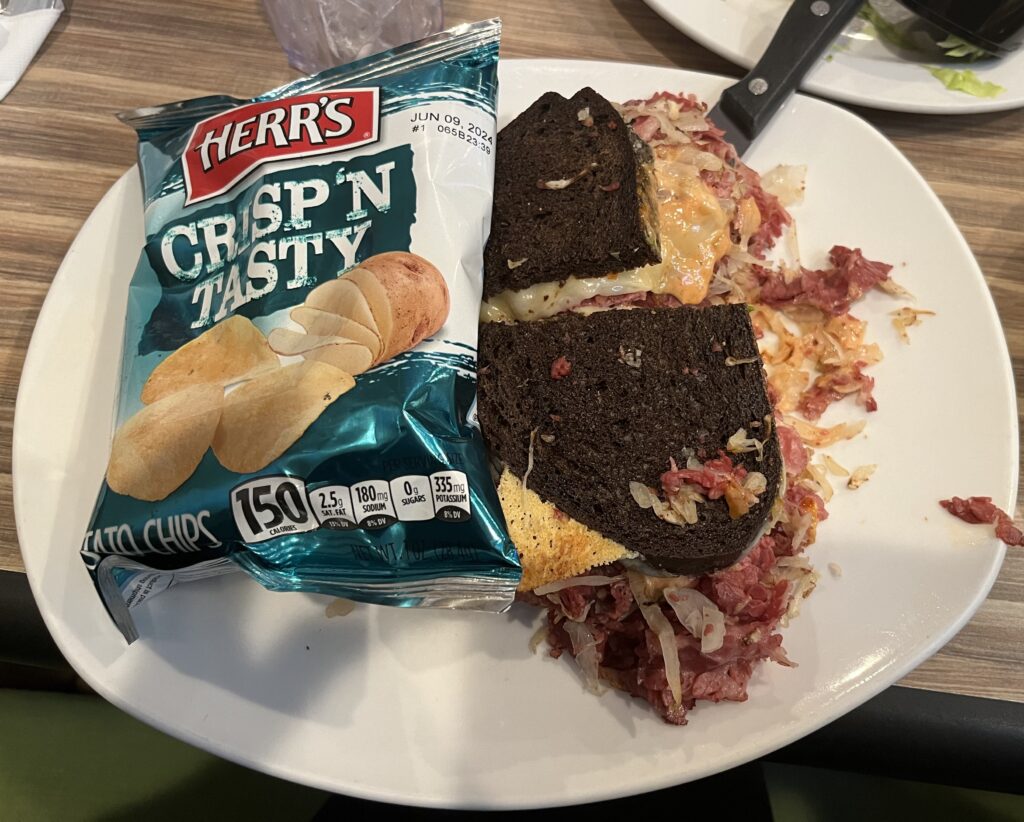 A reuben on dark colored pumpernickle bread served with a bag of chips.