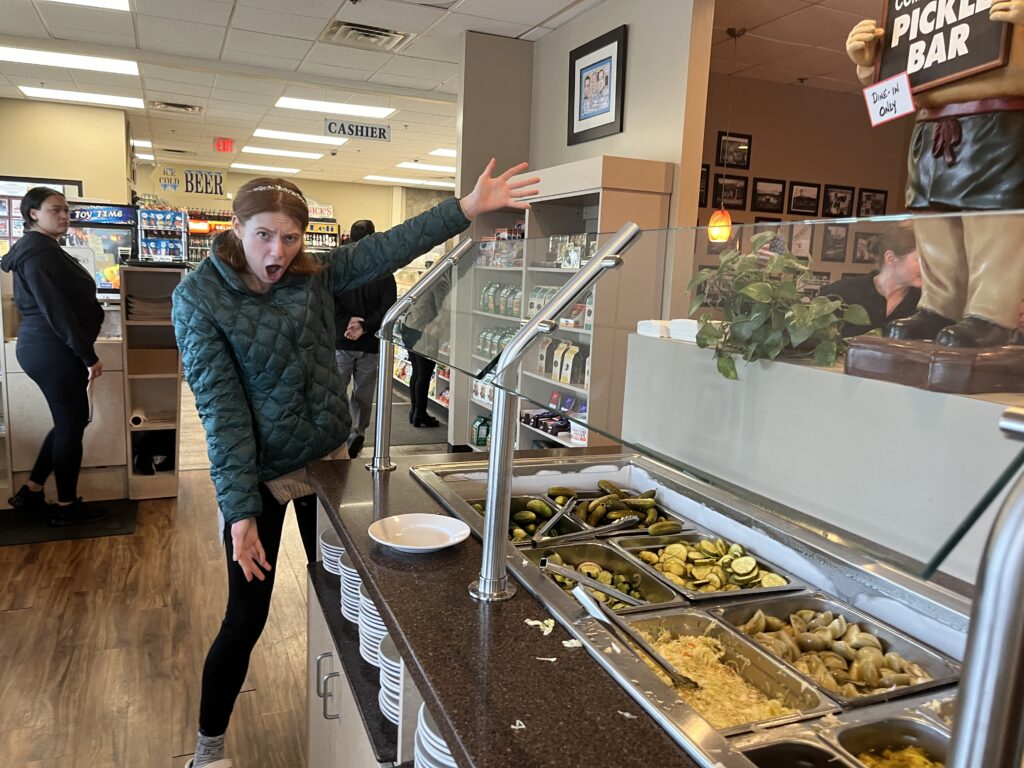 Bri stands excitedly by the picklebar with hands out presenting it.