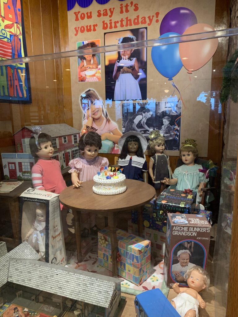 Dolls are posed around a table with a birthday cake on it like at a party.