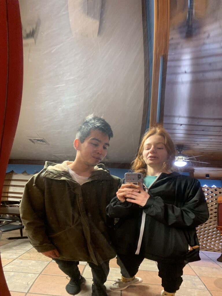 Alix and Bri stand in front of a "fun mirror" making their foreheads tall and body short.