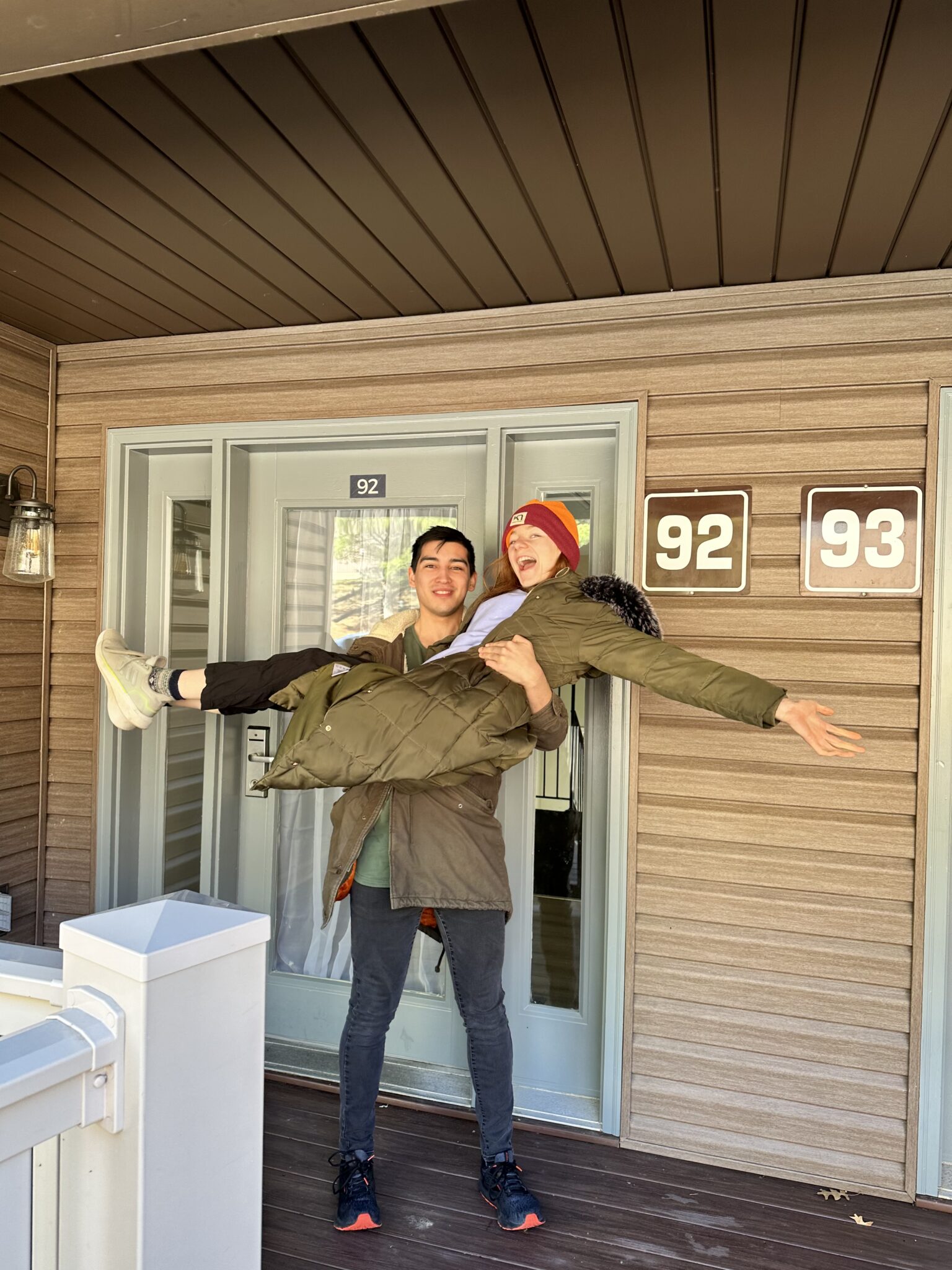 Alix (Bri's boyfriend) holds bri in the classic 'cradle' pose and smiles Bri has her arm out wide smiling in front of the front door of a condo