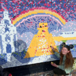 Bri squats near a yellow ziggurat adding her paint swatch to the community art mural.