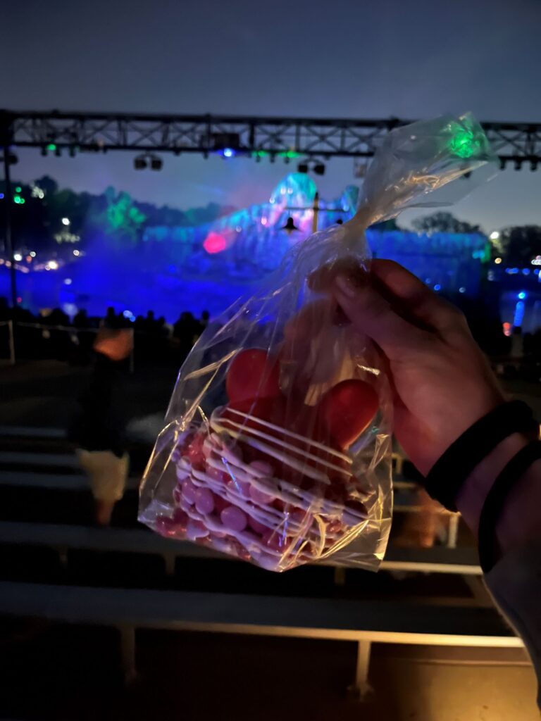 a red chocolate covered mickey shaped candy apple with white chocolate drizzle and valentine color M'n'Ms held in front of the Fantasmic stage,
