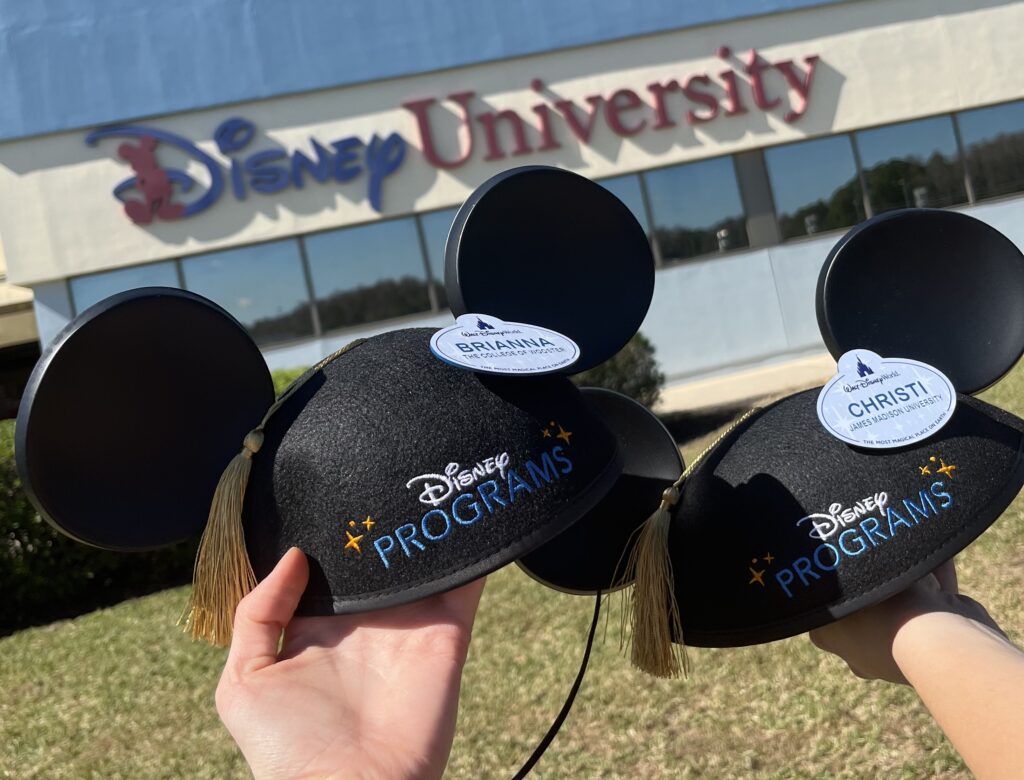 Two mickey ear grad caps one with the cast member name tag "Brianna" the other with the cast member name tag "christi" held in front of a building labeled "Disney University"
