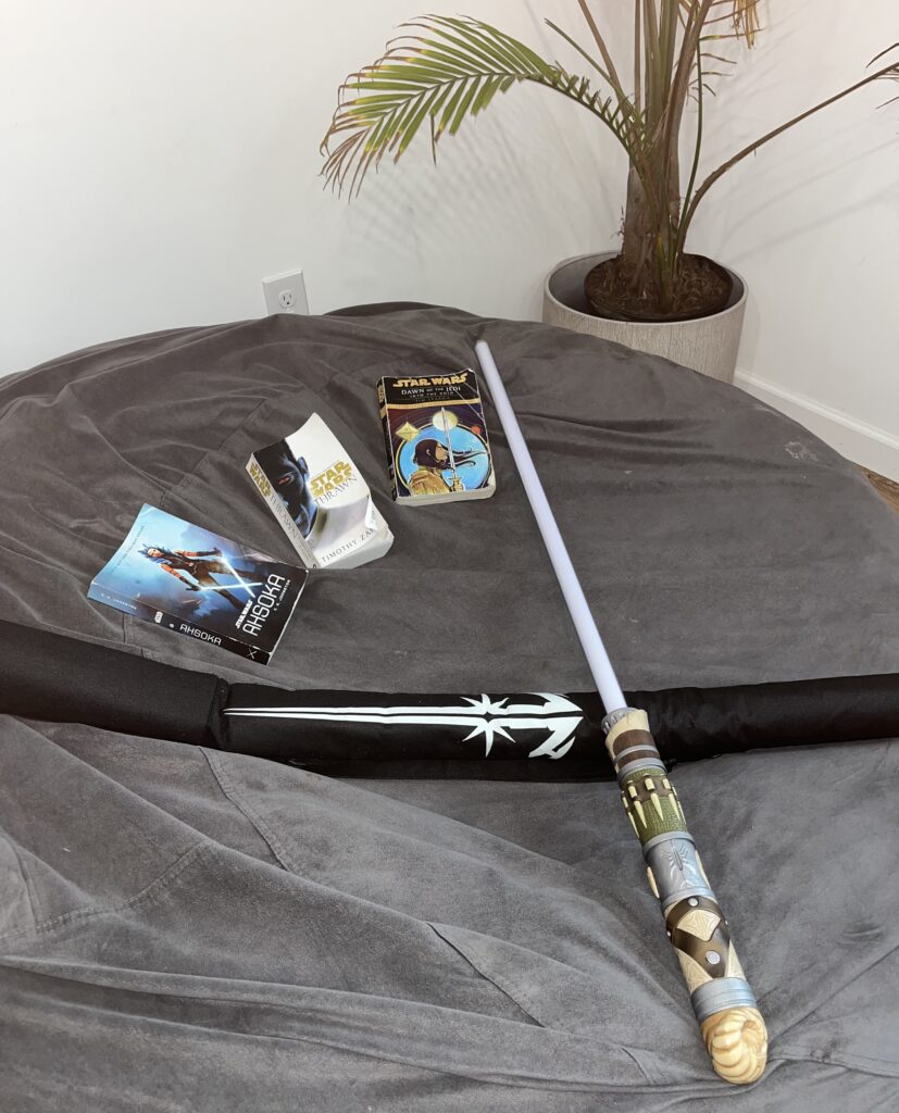 All three Star Wars paperback books laying on a beanbag chair with a lightsaber and lightsaber sheath making an X outside of the books.