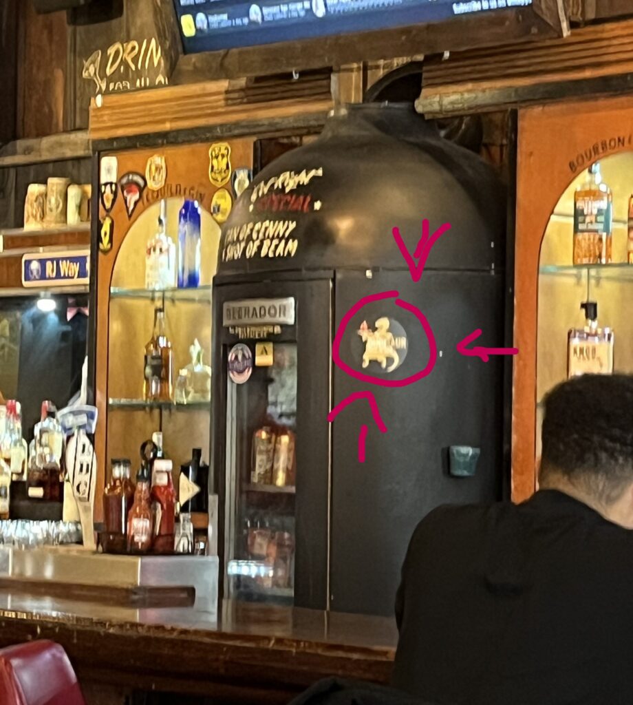 A T-Rex sticker on an old atomic bomb looking shelf holding alcohol at the bar inside Dino BBQ restaurant.