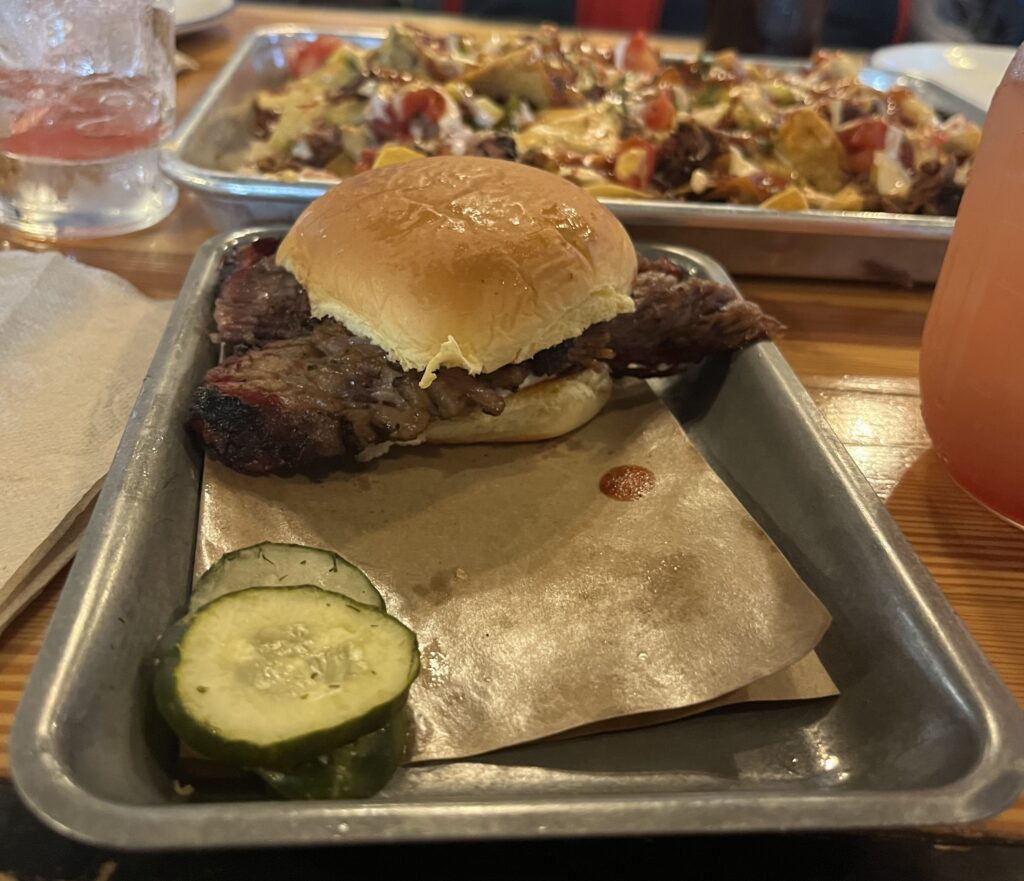 A brisket slider on a small metal tray and pickles on the side.
