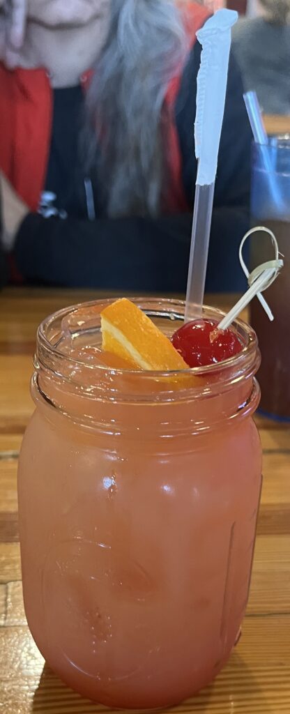 A pink drink in a mason jar with a toothpick skewering a maracchino cherry and orange.