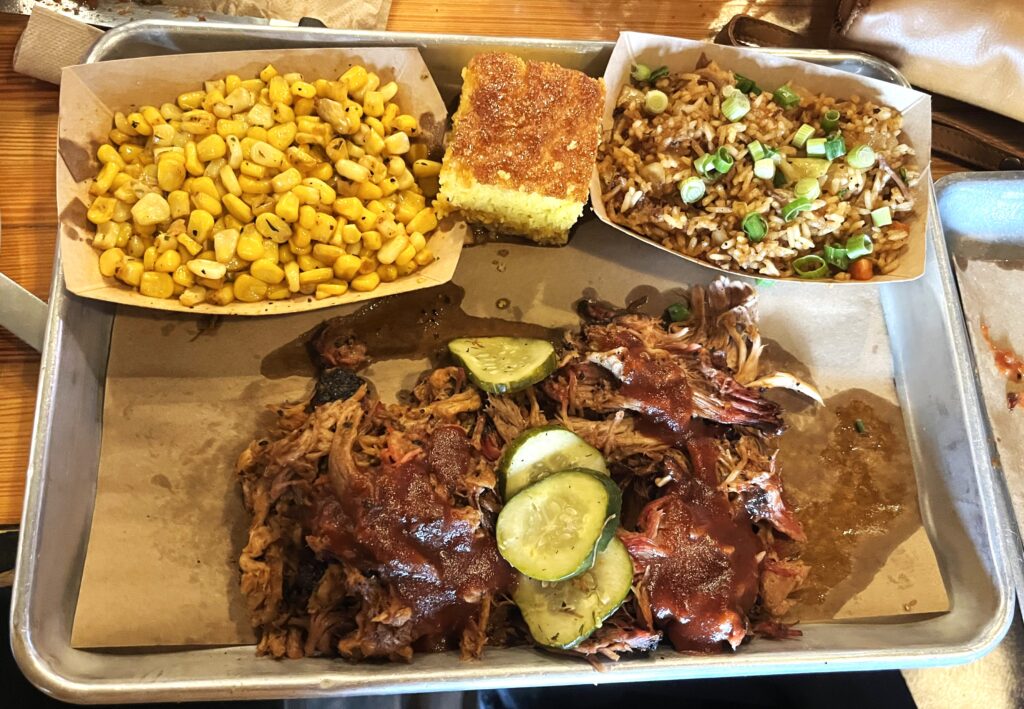 Shredded pork shoulder with BBQ slather and pickles at bottom center of large metal tray, a cardboard tray of corn in the left upper corner, a piecce of corn bread, and a cardboard tray of fried rice in upper right corner.