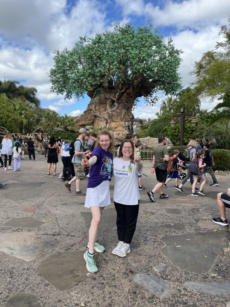 Bri and Sarah stand in front of the Tree of Life (A giant tree with animals carved into it) holding up 1 finger.