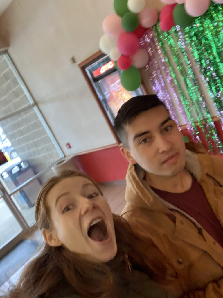 A selfie of Alix and Bri with a pink and green balloon arch and shiny green streamers wall display in the background.
